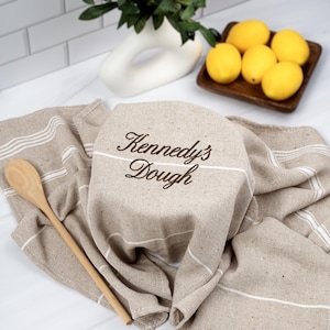 May include: Tan kitchen towel with white stripes and the embroidered text "Kennedy's Dough". A wooden spoon rests on the towel. Lemons in a wooden tray and a white vase with greenery are in the background.