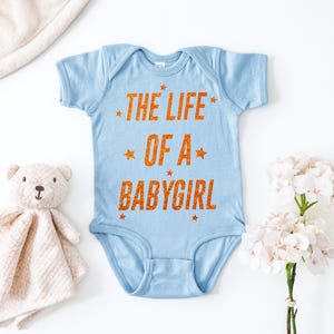 May include: A light blue baby bodysuit with the text "THE LIFE OF A BABYGIRL" in orange. The text is surrounded by orange stars. The bodysuit is made of soft material.