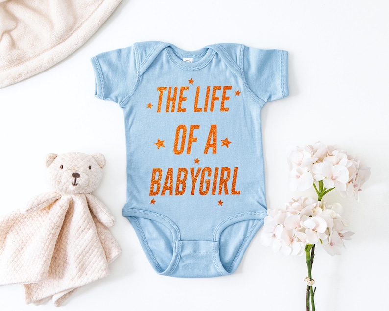 May include: A light blue baby bodysuit with the text "THE LIFE OF A BABYGIRL" in orange. The text is surrounded by orange stars. The bodysuit is made of soft material.
