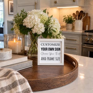 Small white-framed custom sign on a round wooden tray on a kitchen island, beside a vase of white flowers and a candle.