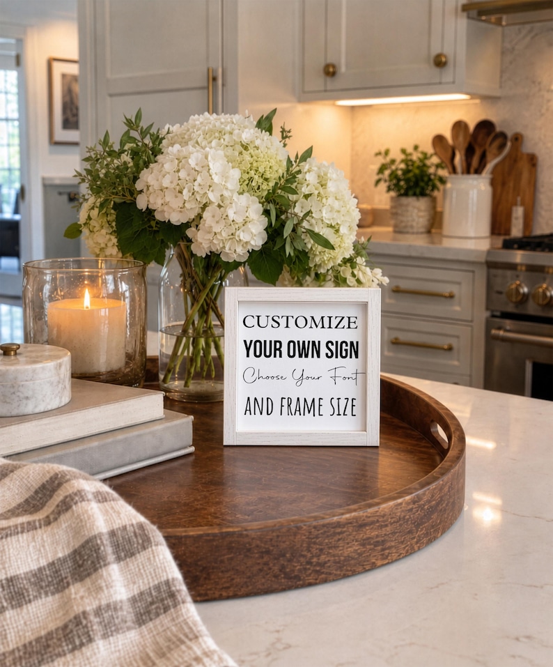 Small white-framed custom sign on a round wooden tray on a kitchen island, beside a vase of white flowers and a candle.