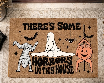 Puede incluir: Un felpudo rectangular de fibra de coco color canela con texto negro que dice THERE'S SOME HORRORS IN THIS HOUSE. El diseño incluye una momia blanca haciendo el pino, un fantasma blanco haciendo un spagat y una calabaza naranja en un poste.