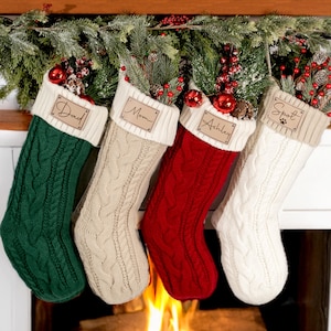 May include: Four cable-knit Christmas stockings in green, beige, red, and white, each with a cream-colored cuff and a wooden name tag. The tags read "Dad", "Mom", "Ashley", and "Spot". Stockings are decorated with greenery and red ornaments.