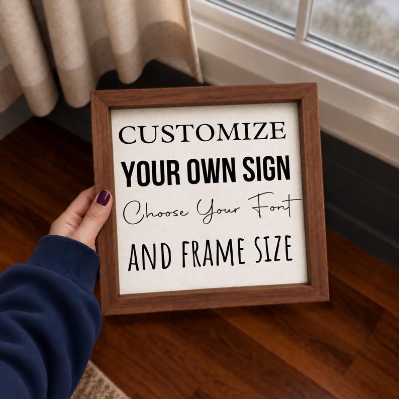 Hand holding a square personalized sign with a brown wood frame beside a window, showing its scale and a sample layout with mixed lettering styles.