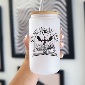 May include: A frosted glass tumbler with a bamboo lid and a clear straw. The glass features a black graphic design of an open book with flowers, a dragon silhouette, moon phases, and the text IN MY FANTASY ERA printed in a serif font.