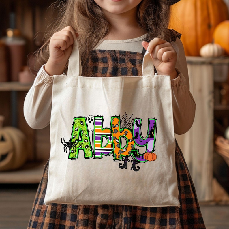 May include: A Halloween-themed tote bag with the name Abby in colorful, decorated letters. The letters are decorated with various Halloween elements such as spiders, ghosts, pumpkins, and bats. The bag is off-white canvas and has a simple design.