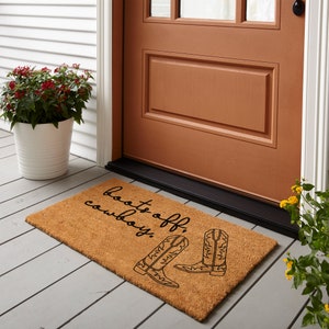 May include: A brown coir doormat with the text "Boots off, cowboy!" in black script. Two black cowboy boots are illustrated on the mat.