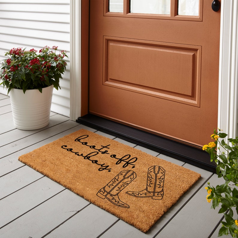 May include: A brown coir doormat with the text "Boots off, cowboy!" in black script. Two black cowboy boots are illustrated on the mat.