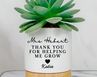 May include: A white cylindrical ceramic planter with a light wood base containing a green succulent. The planter features black text that reads Mrs. Hebert THANK YOU FOR HELPING ME GROW with a small black heart icon above the name Katie.