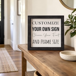 Black-framed custom sign on a wooden entryway console beside a bowl and potted plant. The sample lettering shows different font styles on a white background.