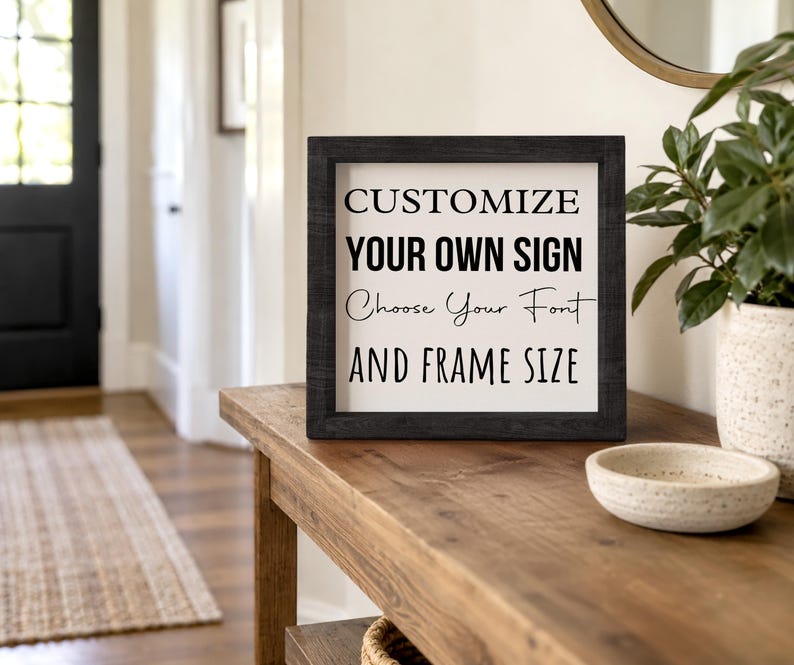 Black-framed custom sign on a wooden entryway console beside a bowl and potted plant. The sample lettering shows different font styles on a white background.