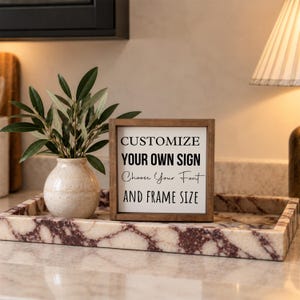 Brown-framed custom wording sign displayed in a rectangular marble tray with a small vase of greenery beside a kitchen lamp.