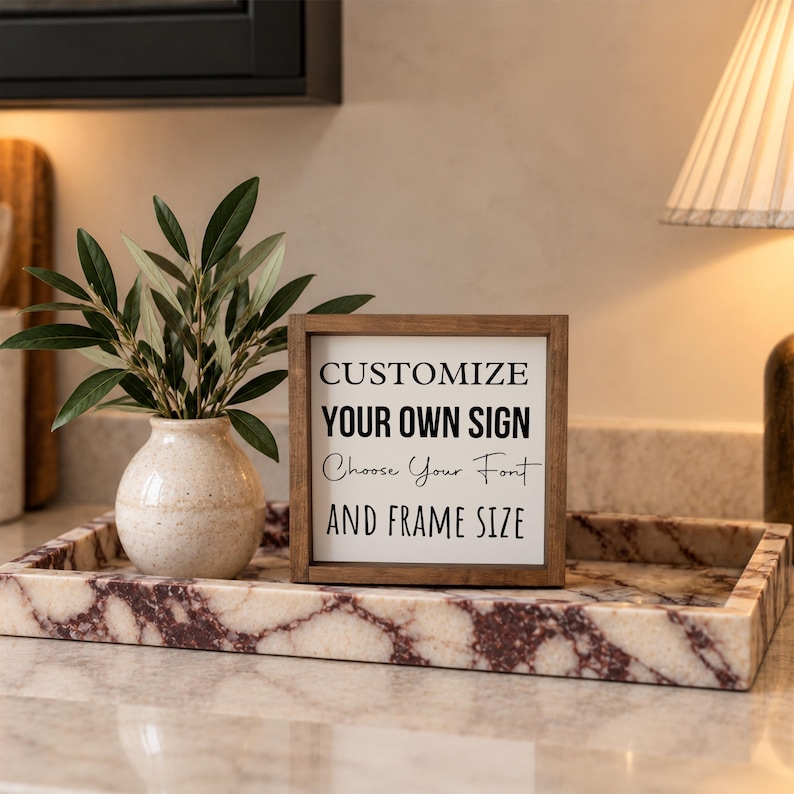 Brown-framed custom wording sign displayed in a rectangular marble tray with a small vase of greenery beside a kitchen lamp.
