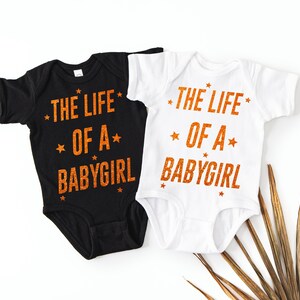 May include: Two baby onesies, one black and one white, with the text "THE LIFE OF A BABYGIRL" in orange glitter lettering. The design includes star accents. A cute and stylish outfit for a baby girl.