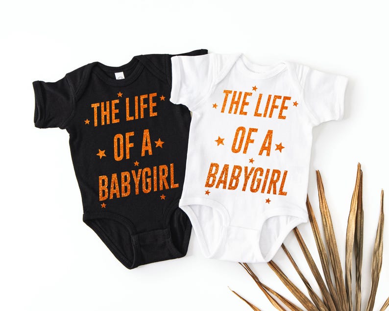 May include: Two baby onesies, one black and one white, with the text "THE LIFE OF A BABYGIRL" in orange glitter lettering. The design includes star accents. A cute and stylish outfit for a baby girl.