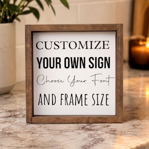 Close view of a brown-framed personalized quote sign on a stone kitchen counter beside a leafy plant, with warm lighting in the background.