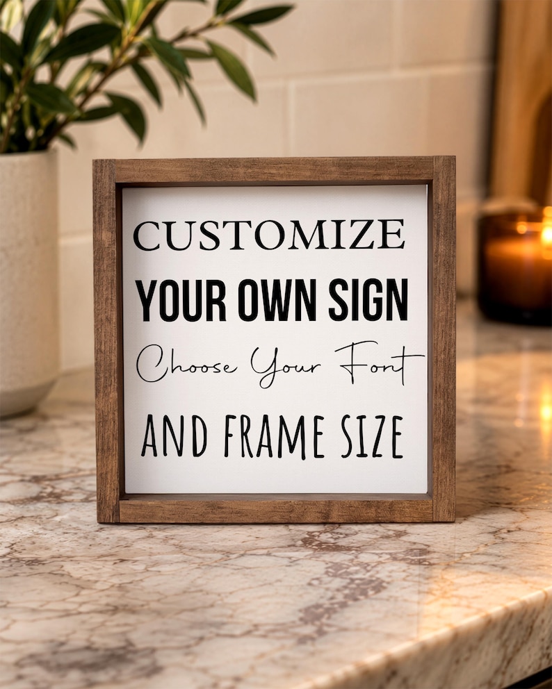 Close view of a brown-framed personalized quote sign on a stone kitchen counter beside a leafy plant, with warm lighting in the background.