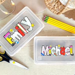 Może przedstawiać: Dwa prostokątne, p&oacute;łprzezroczyste plastikowe pi&oacute;rniki z personalizowanymi imionami nadrukowanymi na wieczkach. Jeden pi&oacute;rnik zawiera imię Emily w kolorowych, szkolnych literach blokowych, a drugi imię Michael w podobnym stylu.