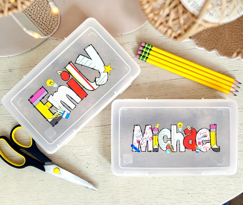 Może przedstawiać: Dwa prostokątne, p&oacute;łprzezroczyste plastikowe pi&oacute;rniki z personalizowanymi imionami nadrukowanymi na wieczkach. Jeden pi&oacute;rnik zawiera imię Emily w kolorowych, szkolnych literach blokowych, a drugi imię Michael w podobnym stylu.