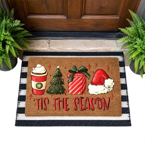 Op de afbeelding: Een rechthoekige bruine kokosmat met een grafische print van een koffiebeker, een kerstboom, een rood cadeau en een kerstmuts. De tekst 'TIS THE SEASON' staat in het rood onder de illustraties, geplaatst op een zwart-wit gestreepte ondergrond.