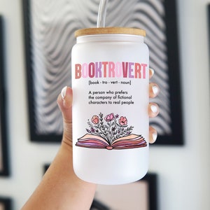 Op de afbeelding: Een matglazen beker met een bamboe deksel en een doorzichtig rietje. De tekst luidt BOOKTROVERT [book-tro-vert-noun] A person who prefers the company of fictional characters to real people, met een illustratie van een open boek en bloemen.