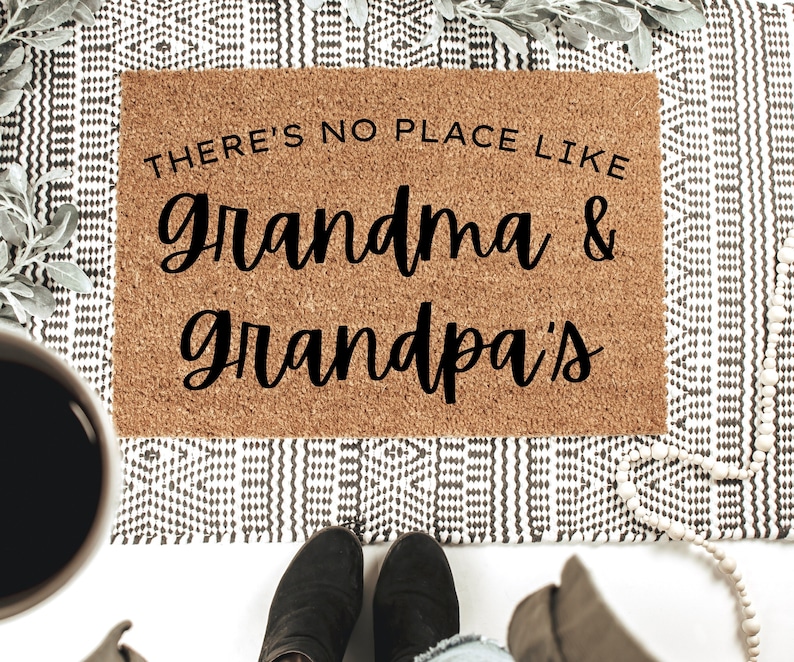 May include: A rectangular coir doormat with the text "THERE'S NO PLACE LIKE grandma & grandpa's" in black script. The mat is placed on a patterned rug. A coffee cup and boots are visible in the frame, adding context to the scene.