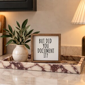 Brown-framed documentation reminder sign on a veined stone tray beside a vase of greenery and a lamp.