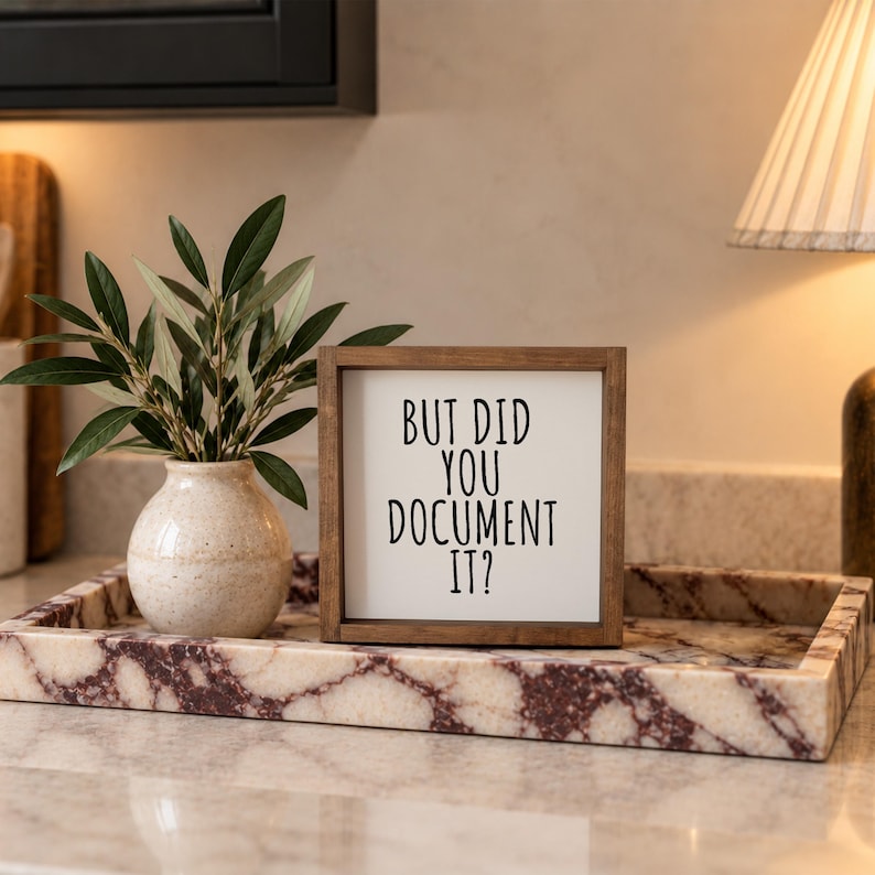Brown-framed documentation reminder sign on a veined stone tray beside a vase of greenery and a lamp.