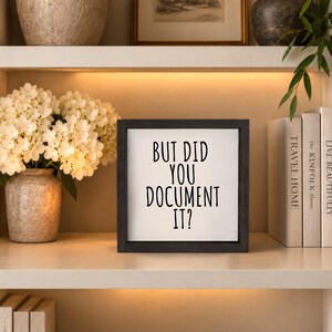 Black-framed “But Did You Document It?” sign on an illuminated shelf beside white flowers and upright books.