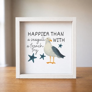 May include: A square white wooden frame containing a print with the text "HAPPIER THAN a seagull WITH a french fry". The illustration features a grey and white seagull with an orange beak and feet, accompanied by three dark blue starfish.