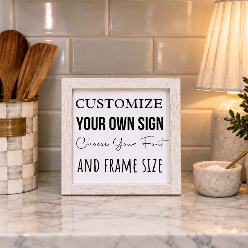 White-framed custom wording sign on a kitchen counter beside a utensil holder and lamp. The sample design invites buyers to choose their font and frame size.