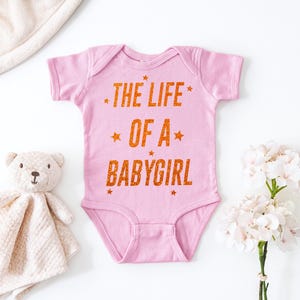 May include: A pink baby onesie with the text "THE LIFE OF A BABYGIRL" in orange glitter lettering. The onesie is surrounded by a stuffed teddy bear, a blanket, and white flowers.