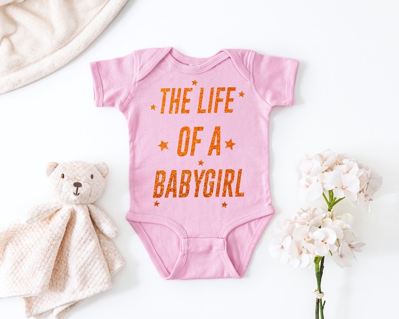 May include: A pink baby onesie with the text "THE LIFE OF A BABYGIRL" in orange glitter lettering. The onesie is surrounded by a stuffed teddy bear, a blanket, and white flowers.