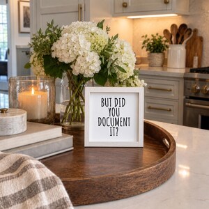 White-framed “But Did You Document It?” sign on a round wooden kitchen tray beside hydrangeas, books, a candle, and a checked cloth.