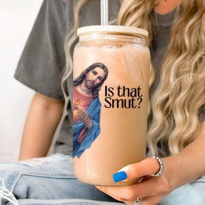 May include: A clear glass tumbler with a bamboo lid and a straw, filled with a light brown beverage. The tumbler features an image of Jesus and the text "Is that Smut?" in black. The tumbler is held by a person.