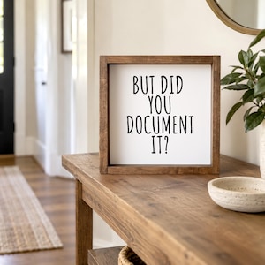 Brown-framed “But Did You Document It?” sign on a wooden entryway console beside a leafy plant and shallow bowl. Tall black lettering is arranged on a light background.