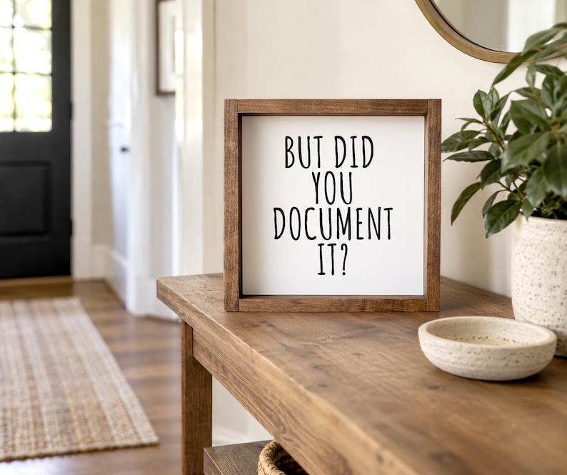Brown-framed “But Did You Document It?” sign on a wooden entryway console beside a leafy plant and shallow bowl. Tall black lettering is arranged on a light background.