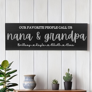 May include: A rectangular black wooden sign with white text that reads OUR FAVORITE PEOPLE CALL US nana & grandpa, followed by the names Brittany, Kayla, Elliott, and Elena separated by small heart icons in a white script font.