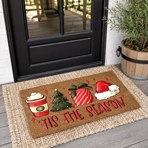 Op de afbeelding: Een rechthoekige bruine kokosmat met een rode koffiebeker, een groene kerstboom, een rood cadeau met een groene strik en een rode kerstmuts. De tekst 'TIS THE SEASON' staat in rode letters onderaan de deurmat afgebeeld.
