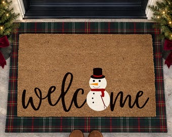 May include: A rectangular brown coir doormat featuring the word welcome in black cursive script. A white snowman with a black top hat, orange carrot nose, and red scarf is positioned in the center, replacing the letter o in welcome.