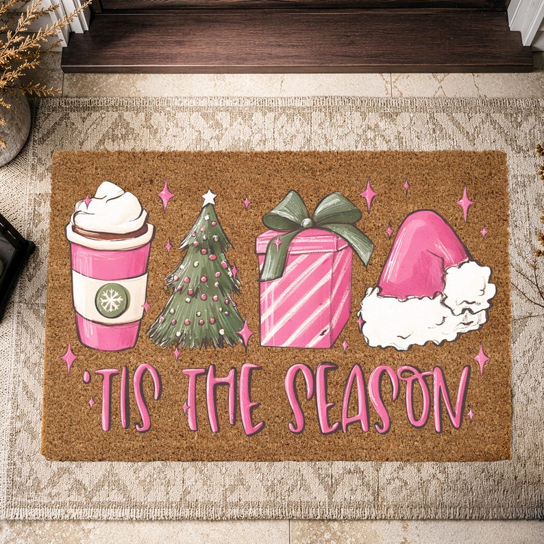 Op de afbeelding: Een rechthoekige bruine kokosmat met een roze koffiebeker, een groene kerstboom, een roze gestreept cadeau met een groene strik en een roze kerstmuts. De tekst 'TIS THE SEASON' staat in roze letters onderaan de mat gedrukt.