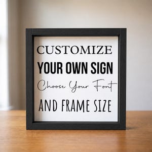 Straight-on view of a black wood frame on a wooden table, displaying the 'Customize your own sign' sample with serif, script, and block lettering.