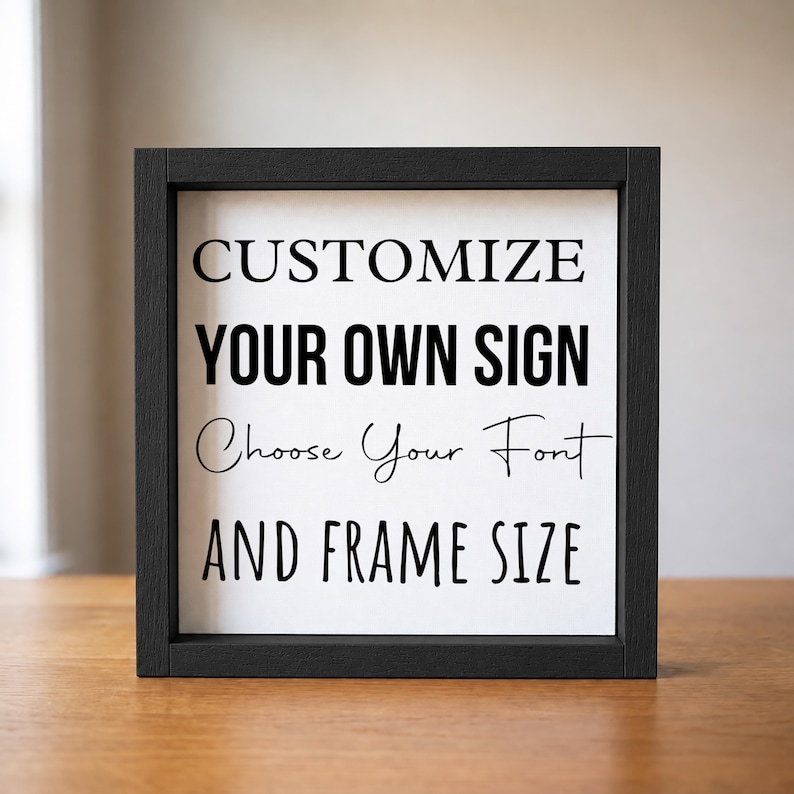 Straight-on view of a black wood frame on a wooden table, displaying the 'Customize your own sign' sample with serif, script, and block lettering.