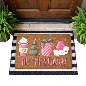 Op de afbeelding: Een rechthoekige bruine kokosmat met een grafische print van een roze koffiebeker, een groene kerstboom, een roze cadeau met een groene strik en een roze kerstmuts. De tekst 'TIS THE SEASON' staat in roze letters onder de afbeeldingen.