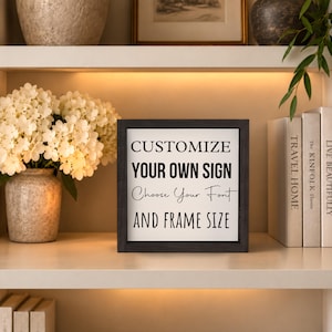 Black-framed custom quote sign displayed on a softly lit bookshelf between flowers and books, with black sample lettering on a white background.