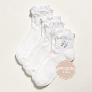 K&ouml;nnte beinhalten: Vier wei&szlig;e Kn&ouml;chelsocken mit R&uuml;schen. Jede Socke hat einen hellblau gestickten Buchstaben oder Initialen. Die Socken werden auf wei&szlig;em Hintergrund pr&auml;sentiert. Ein hellrosa Aufkleber mit den Worten "EMBROIDERED DESIGN" befindet sich in der unteren rechten Ecke.