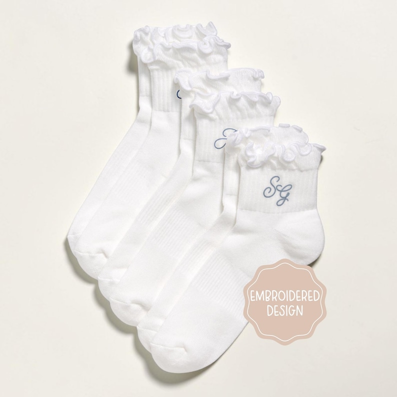 K&ouml;nnte beinhalten: Vier wei&szlig;e Kn&ouml;chelsocken mit R&uuml;schen. Jede Socke hat einen hellblau gestickten Buchstaben oder Initialen. Die Socken werden auf wei&szlig;em Hintergrund pr&auml;sentiert. Ein hellrosa Aufkleber mit den Worten "EMBROIDERED DESIGN" befindet sich in der unteren rechten Ecke.