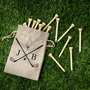 May include: A beige burlap drawstring bag with a golf-themed design, including crossed golf clubs, a golf ball, and the initials "JB". The bag is filled with wooden golf tees, each marked with "JB", scattered on a green grass background.
