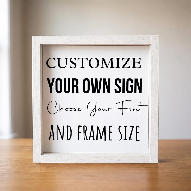 Front view of a white wood frame surrounding the black-and-white 'Customize your own sign' sample, standing on a wooden table against a light wall.