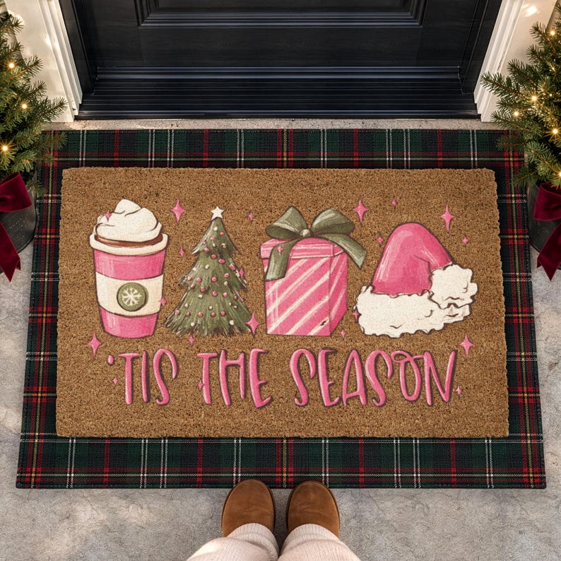 Op de afbeelding: Een rechthoekige bruine kokosmat met een roze koffiebeker met slagroom, een groene kerstboom, een roze gestreept cadeau met een groene strik en een roze kerstmuts. De tekst 'TIS THE SEASON' is in roze letters op de mat gedrukt.